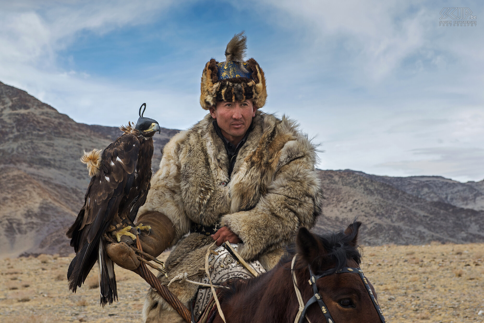 Ulgii - Golden Eagle Festival - Berkutchi A Kazakh eagle hunter is called kusbegi/kushbegi/qusbegi or berkutchi. In Turkic languages kus(h)begi means ‘bird lord’ or ‘bird master’. Berkut is the word for ‘golden eagle’ and ‘chi’ is the end tag for words. The berkutchi mostly wear their traditional costumes. Their fur coats are made of marmot, fox or wolf skins caught by their eagles. The eagle hunters always wear a 'tomach', a fur cap of fox skin combined with red, fuschia or blue fabric. The more extravagant the coat, the more respected the hunter. Stefan Cruysberghs